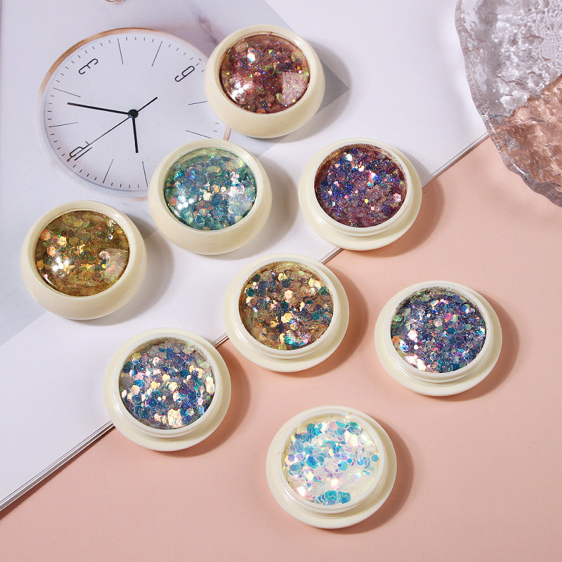 Eight small, round nail glitter sequin decals in various colors placed on a surface with a clock and other decorative items in the background.